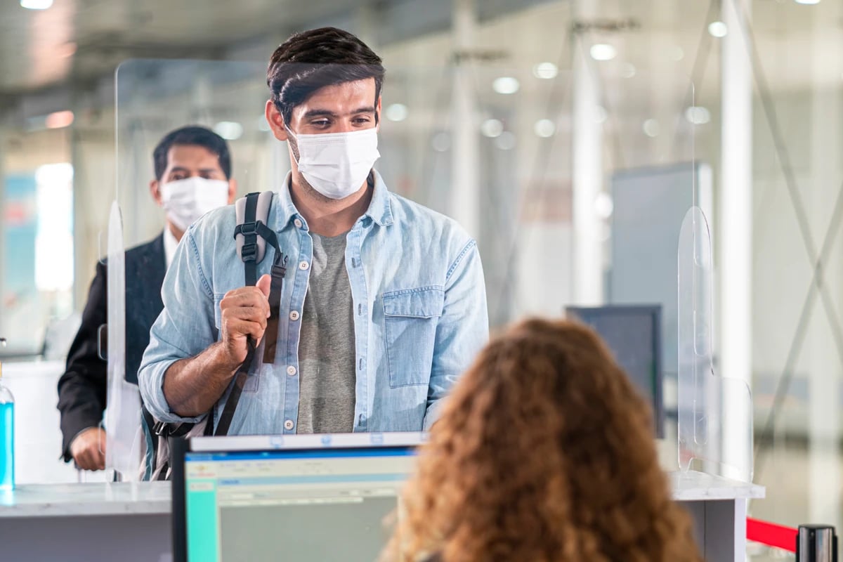 airline-passenger-behind-clear-panel-wearing-mask-conversating-with-airline-employee-sitting-at-desk-with-computer